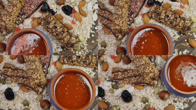 顶视图。陶罐中的新鲜蜂蜜，以谷物能量棒和燕麦片为背景，配以整个坚果，浆果和种子。旋转，特写。