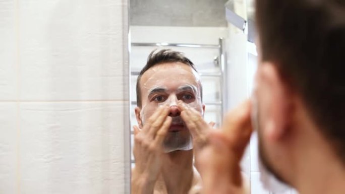 镜子反射微笑千禧一代男人在浴室里涂化妆品面膜。在家进行自我护理、健康和水疗护理。