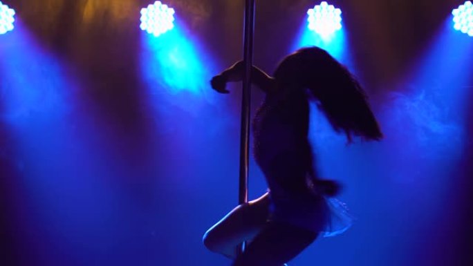 在一个有蓝色霓虹灯的黑暗工作室里，一个运动灵活的女人在一根杆子上旋转。舞蹈飞行元素编舞，健美操，健身