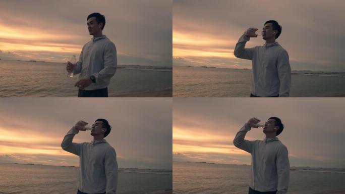亚洲运动员运动员站在海滩上喝水美丽的日落。运动员训练运动锻炼保健概念。