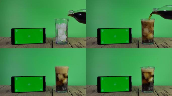 将可口可乐倒入绿色背景的绿色屏幕的玻璃和智能手机中。