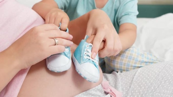 小男孩在怀孕的母亲大肚子上玩婴儿鞋的特写镜头。开朗的孩子与期待宝宝的家庭幸福的概念