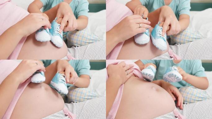 小男孩在怀孕的母亲大肚子上玩婴儿鞋的特写镜头。开朗的孩子与期待宝宝的家庭幸福的概念
