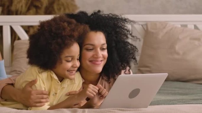非裔美国妇女和女孩在笔记本电脑上打字，浏览内容和交流。母亲和女儿躺在卧室的床上，室内明亮。特写。慢动