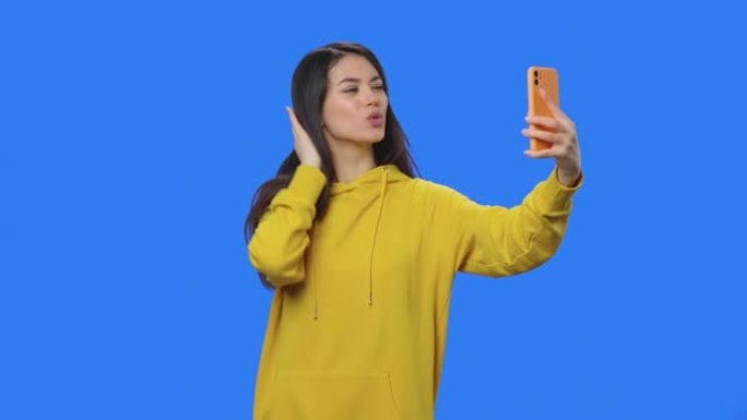 黑发女人的肖像在手机上自拍，做有趣的脸，看照片。穿着黄色运动衫的年轻女子在蓝屏背景的工作室摆姿势。特