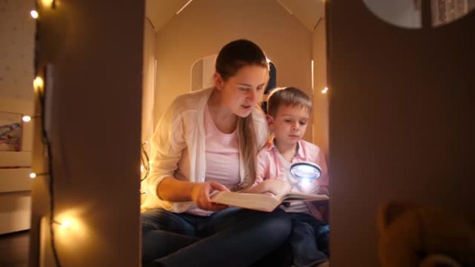 多莉 (Dolly) 拍摄的母亲和小儿子在晚上在由花环照亮的小玩具屋中读童话书的镜头。儿童教育和家庭