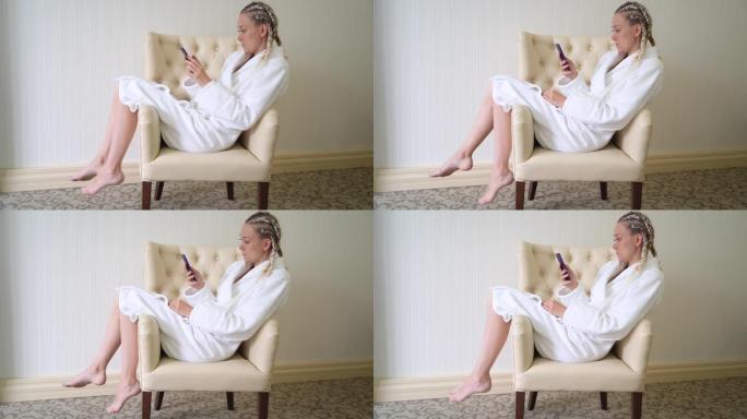 穿着白色浴袍的年轻女子坐在扶手椅上时使用手机。
