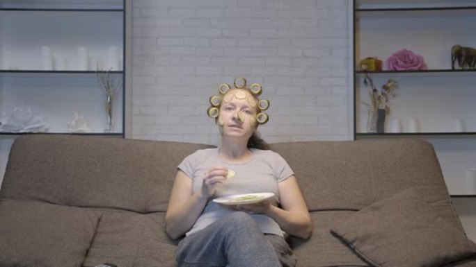 戴面膜的成年妇女坐在沙发上看电视时吃黄瓜