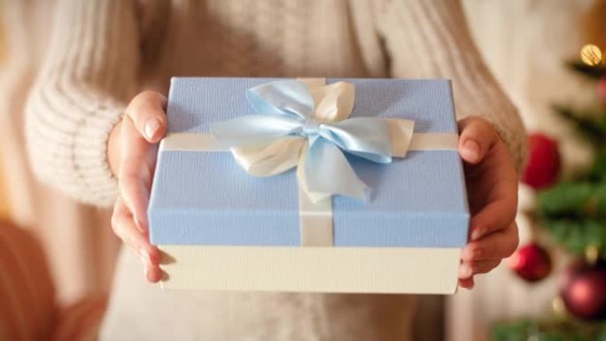 特写4k镜头，年轻女子拿着漂亮的盒子，礼物用丝带和蝴蝶结绑着。家庭在圣诞树旁边的客厅赠送和接收新年礼