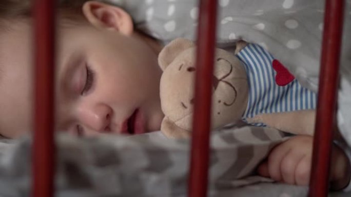 关闭正宗可爱的小婴儿胖乎乎的女婴或男孩在午餐时间甜蜜地睡在卧室和泰迪熊的婴儿床里。孩子躺在灰色的床上