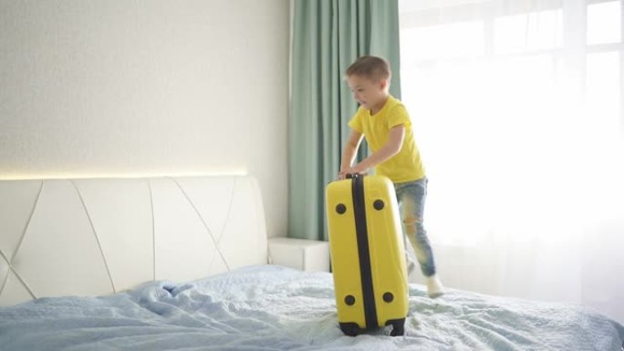 头发浅的漂亮男孩在旅途中很开心，微笑着。一个带着黄色手提箱的孩子在旅馆的床上跳起来欢喜
