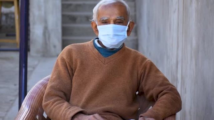 戴着面具盯着相机的印度老人的肖像。新型冠状病毒肺炎的老年护理。坐在室内保护自己免受电晕病毒大流行的高