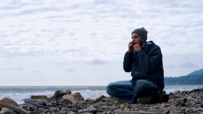 留着白种人胡子的男孩坐在海浪的背景下，坐在多岩石的海岸上，用嘴创作音乐，享受生活。