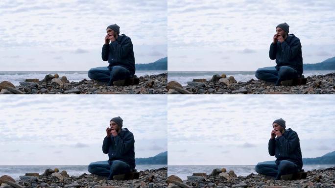 留着白种人胡子的男孩坐在海浪的背景下，坐在多岩石的海岸上，用嘴创作音乐，享受生活。
