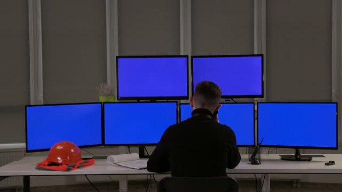 一位工程师坐在许多电脑屏幕前。桌子上是橙色的头盔和背心。从后面看。他在打电话。