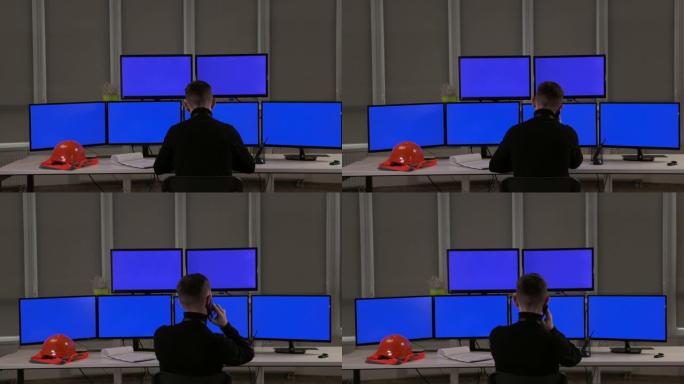 一位工程师坐在许多电脑屏幕前。桌子上是橙色的头盔和背心。从后面看。他在打电话。