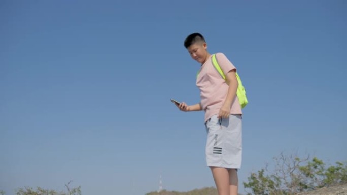 亚洲背包客男孩在蓝天的山顶上通过智能手机拍照，生活方式概念。