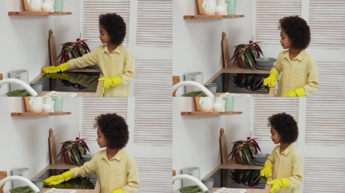小非洲裔美国女孩戴着黄色橡胶手套，用毛巾擦拭电炉表面。十几岁的女孩在明亮的厨房背景下摆姿势。慢动作
