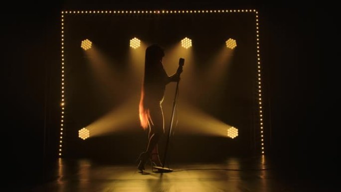 迷人的女歌手剪影在黑暗的舞台上表演个人音乐会，背景是明亮的灯光。穿着闪亮连衣裙的女人触摸老式麦克风，