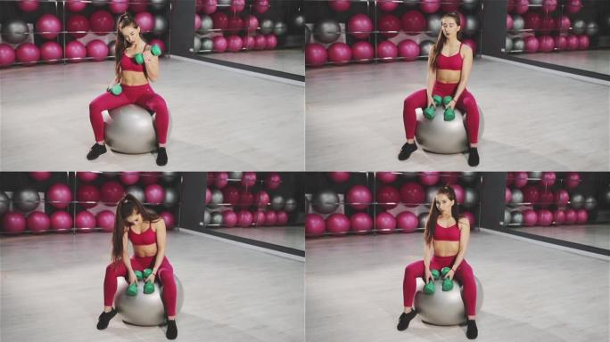 年轻女子坐在健身房的健身球上用哑铃训练