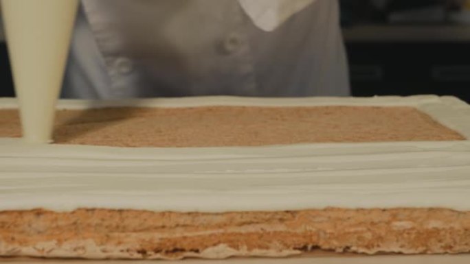 糕点厨师将黄油奶油从冷却器中挤出到海绵蛋糕上，制成酥皮卷。糕点厨师迅速均匀地将奶油涂在蛋糕上。专业工