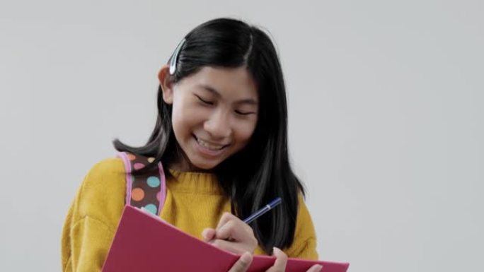 亚洲背包女学生穿着黄色毛衣写在粉红色的书上，在灰色的背景下，生活方式的概念。