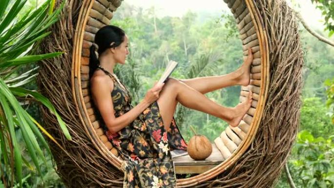 一本书的美丽女孩坐在藤本植物制成的热带椅子上。配椰子