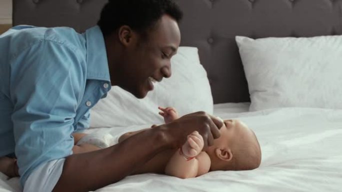 陪产假。快乐的非洲裔美国男子和他可爱的新生婴儿躺在大床上玩耍，微笑着大笑