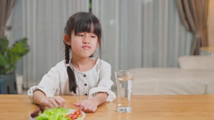 不快乐的亚洲小女孩孩子不想在餐盘上吃绿色蔬菜。小女儿闻到黄瓜的味道，用无聊的脸推开盘子。健康食品和儿