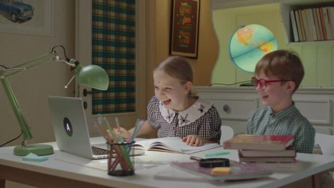两个兄弟姐妹坐在家里的办公桌上在线上课。小学生在家上学期间取笑。兄妹笑着看着笔记本电脑。
