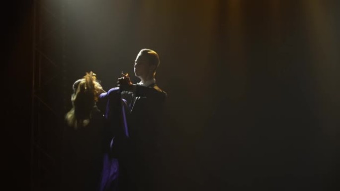 一对穿着优雅服装的浪漫情侣正在跳华尔兹舞。舞厅舞者的剪影在黑暗的烟熏工作室里，在一束舞台灯光下跳舞。