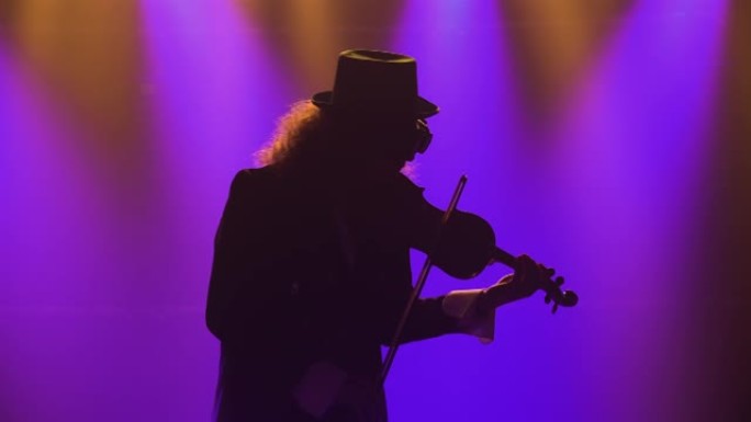 一个穿着复古西装，帽子和原始眼镜的男人拉小提琴。音乐家在黑烟的工作室里用蓝灯表演古典音乐会。剪影。特