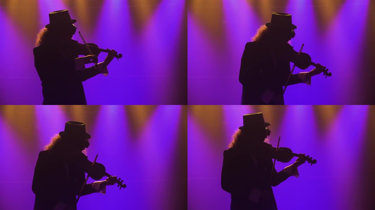 一个穿着复古西装，帽子和原始眼镜的男人拉小提琴。音乐家在黑烟的工作室里用蓝灯表演古典音乐会。剪影。特