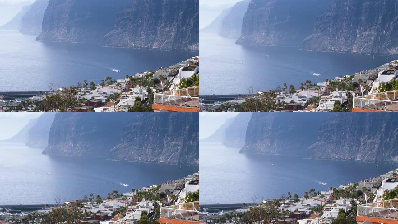 4k视频美丽的白色摩托艇在高高的大悬崖和群山环绕的港口骑行。加那利群岛特内里费洛斯·吉甘特斯的全景景