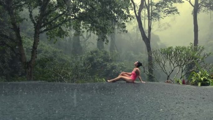 穿着粉色比基尼的美丽年轻女子享受热带雨水落在她身上，在无限泳池中欣赏神秘的丛林景色