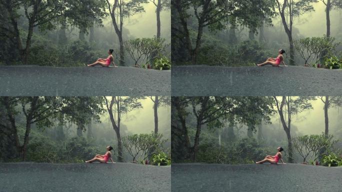 穿着粉色比基尼的美丽年轻女子享受热带雨水落在她身上，在无限泳池中欣赏神秘的丛林景色