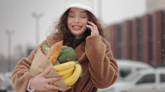 一个女人正在超市附近，用智能手机聊天。她微笑着拿着一个购物袋。大雪正在下。4K。