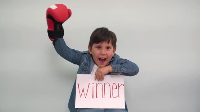 一个戴着拳击手套的男孩拿着一张纸，上面写着题词获胜者，并欢呼一个快乐的孩子在灰蓝色背景上展示了一个带
