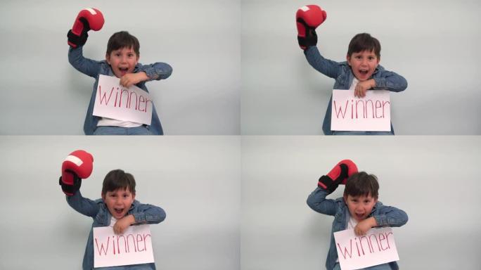 一个戴着拳击手套的男孩拿着一张纸，上面写着题词获胜者，并欢呼一个快乐的孩子在灰蓝色背景上展示了一个带