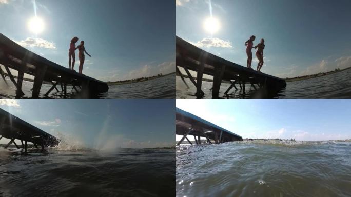在一个阳光明媚的夏日，在蓝天和阳光的背景下，两个快乐的年轻女孩手牵手从码头跳入水中。从下面看，从水位