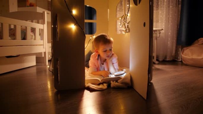 快乐的微笑男孩躺在居屋玩具屋或帐篷的地板上，晚上用手电筒看书。儿童教育与暗室阅读的概念