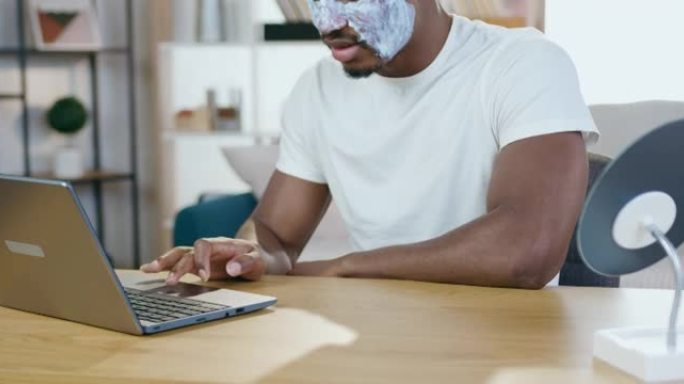 英俊微笑的现代年轻黑皮肤男人，戴着白色面膜，坐在笔记本电脑前，重温有趣的视频，慢动作