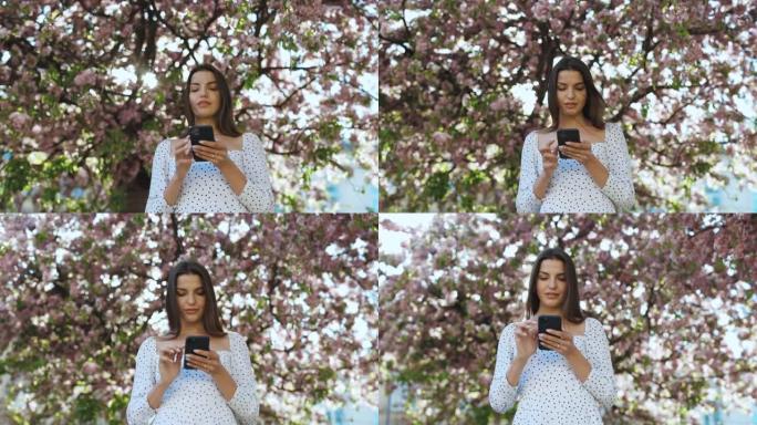 在樱花树的背景下，在公园里使用智能手机的美女肖像。年轻漂亮的女孩使用社交网络浏览互联网