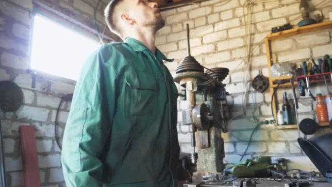 工作服中的男性机械师脱下防护眼镜，擦去额头上的汗水。年轻的修理工戴上眼镜，继续在车库工作。维护服务的