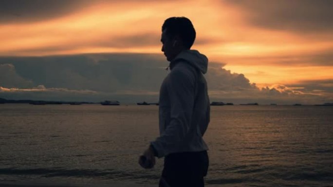 夏天美丽的日落时，亚洲跑步男子在海滩上使用跳绳在户外锻炼。健康运动生活方式理念。