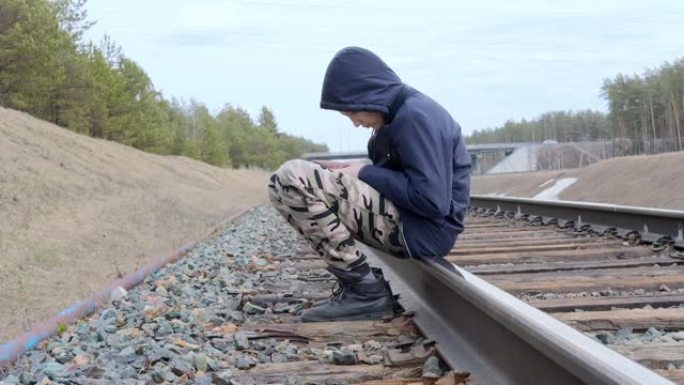 悲伤孤独的少年坐在危险中的铁轨上。男孩歪着头，玩电话，没有注意到周围的任何东西。逃离家庭概念