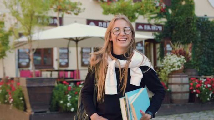 迷人的洋溢着笑容的年轻金发碧眼的女大学生穿着时髦的眼镜，戴着时髦的眼镜，手里拿着书，在学习的路上，在