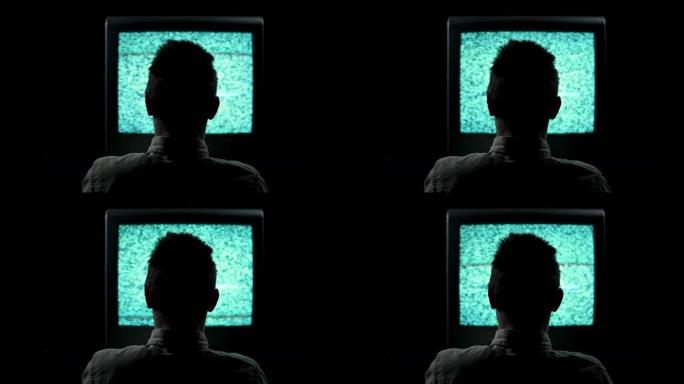 在黑色背景的黑暗演播室里，一个男人坐在闪烁的电视前的轮廓的后视图。一名男子将手撞在一台破损的电视上，
