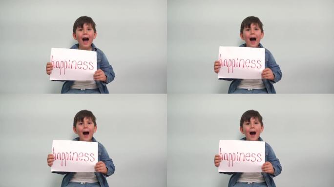 一个男孩手里拿着一张纸，上面写着幸福，张开嘴。一个开朗的孩子在灰蓝色背景上显示了一个带有幸福字样的标