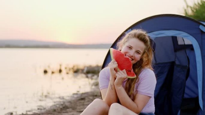 带着一大块西瓜微笑的大女士在日落时在湖边野餐，她享受着时光。在阿里·阿列克谢拍摄